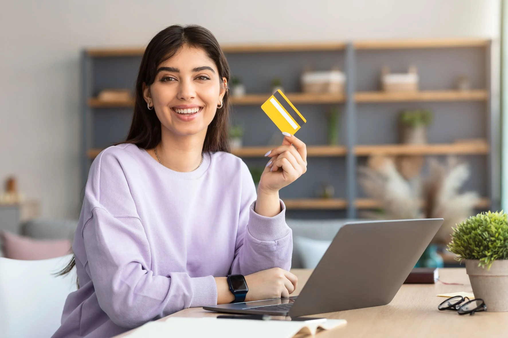 Smiling woman holding a yellow gift card while working on a laptop at home
