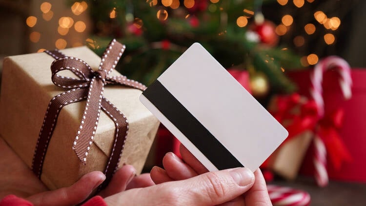 Hand holding a gift card beside a wrapped present and a decorated holiday tree