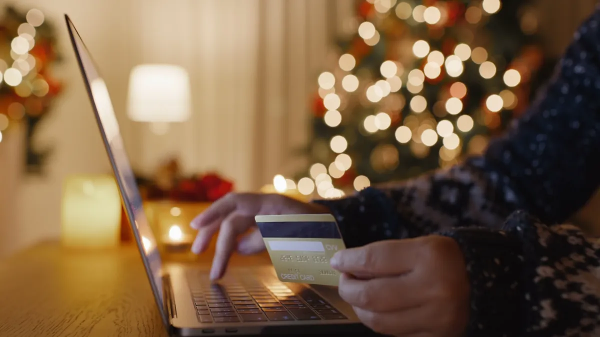 Person holding a card while shopping online on a laptop beside a lit holiday tree